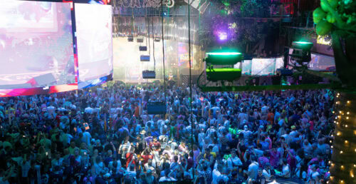 Vor dem Hintergrund eines Nachtclubs oder Konzertsaals mit bunten Lichtern, Bildschirmen und einer Bühne versammelt sich eine große Menschenmenge. Die Menschen blicken nach vorne und verfolgen eine Darbietung.