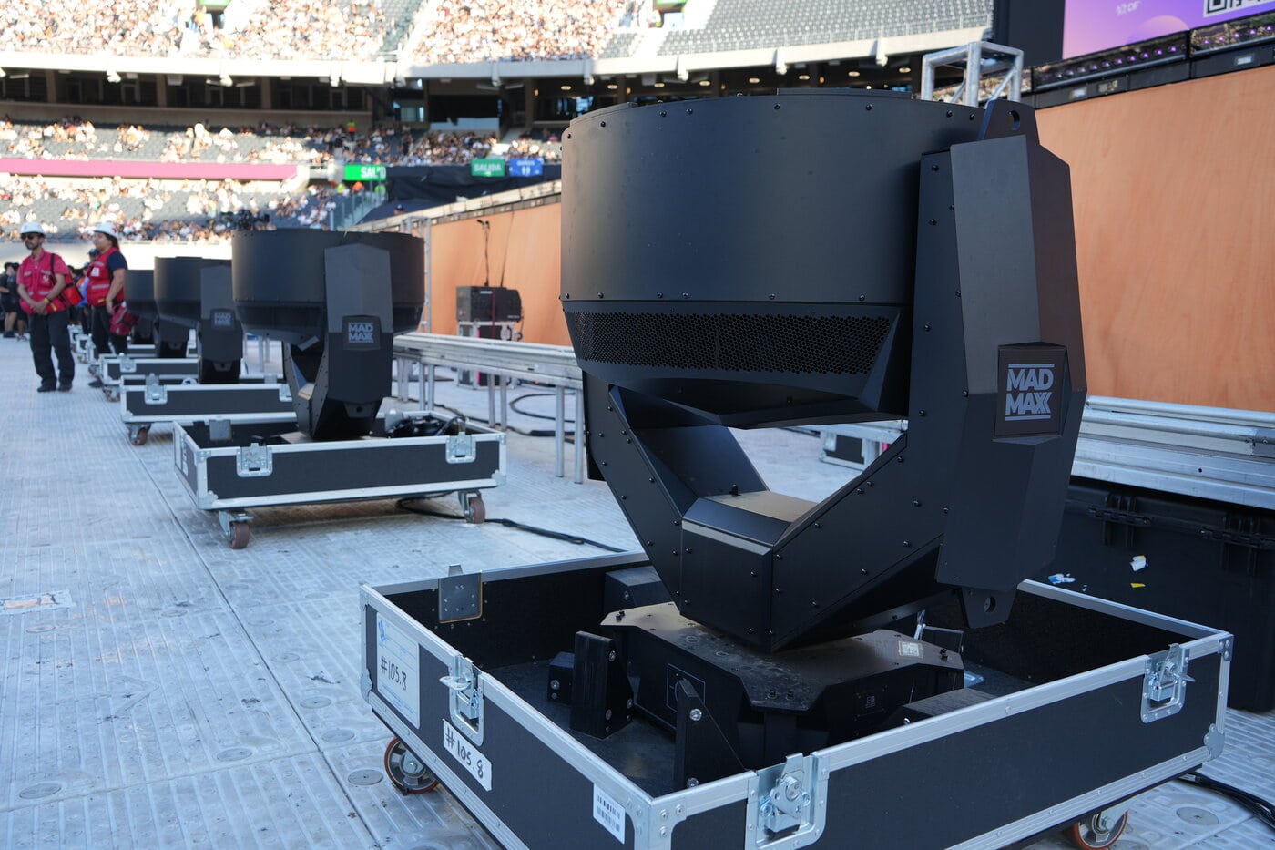 Photo 3 – Credit – Evangelina Bertelllo Mehrere große Bühnenleuchten mit der Aufschrift „Mad Max“ stehen auf Kisten am Rand eines Freiluftstadions, im Hintergrund sind Mitarbeiter der Veranstaltung zu sehen.