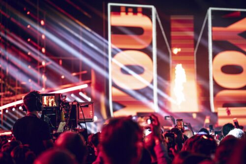 Eine Menschenmenge bei einer Live-Veranstaltung oder einem Konzert mit roter Beleuchtung, großen Bildschirmen, auf denen auffällige Grafiken zu sehen sind, und einem Kameramann, der die Bühne filmt.