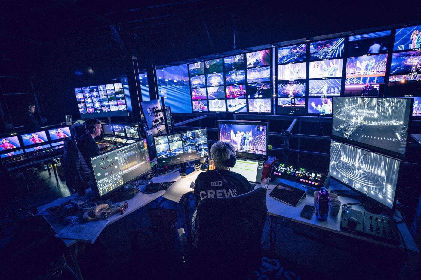 Ein Regieraum für Fernsehproduktionen mit mehreren Bildschirmen, auf denen Live-Übertragungen von Veranstaltungen zu sehen sind. Die Mitarbeiter sitzen an ihren Schreibtischen, bedienen die Technik und überwachen die Sendungen.