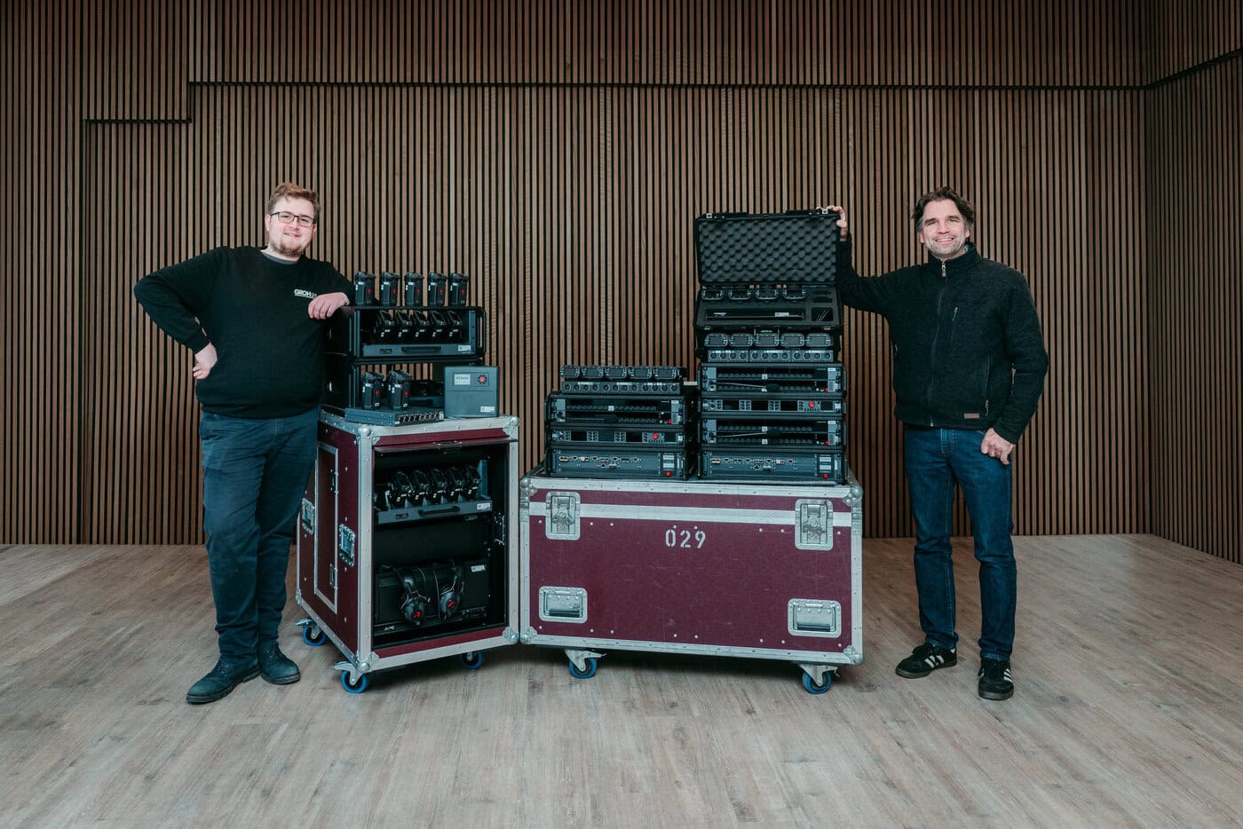 Riedel – Links Fynn Heidutzek – Rechts Jan Grohmann-Falke Zwei Personen stehen in einem Raum mit holzvertäfelten Wänden und posieren neben großen Kisten, die mit elektronischen Geräten und drahtlosen Headset-Systemen auf Rollwagen gefüllt sind.