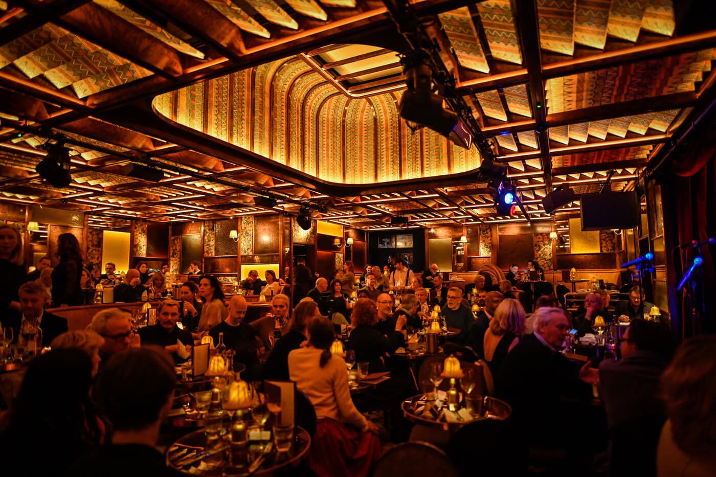 Ein warm beleuchtetes, prunkvoll eingerichtetes Restaurant ist voller Menschen, die an Tischen sitzen, sich unterhalten und essen. Die Decke ist mit geometrischen Mustern verziert, und es gibt eine Bühne mit Mikrofonen.