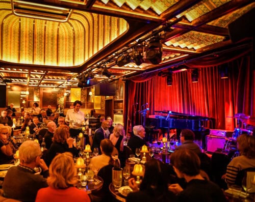 In einem warm beleuchteten Jazzclub mit einer Bühne mit roten Vorhängen, einem Klavier und Instrumenten sitzt eine Gruppe von Menschen an Tischen; ein Kellner bedient die Gäste, und die Leute essen und unterhalten sich.