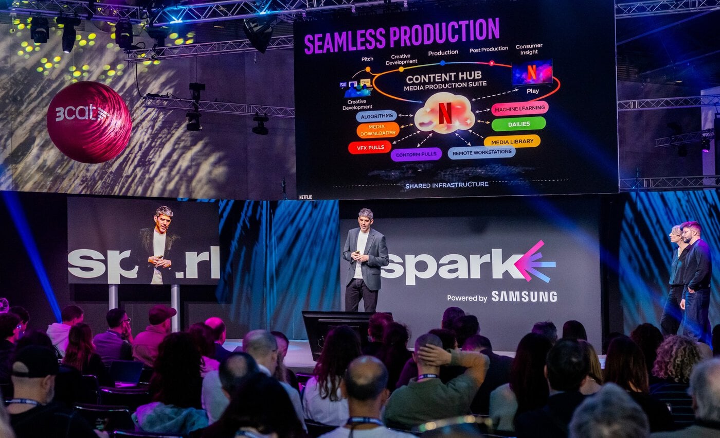 ise-spark Ein Moderator steht bei einer Technologieveranstaltung auf der Bühne und spricht zu den Zuhörern. Auf einer großen Leinwand hinter ihm ist ein Diagramm der „Seamless Production“-Medien-Suite zu sehen.