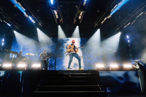 Eine Band tritt auf der Bühne auf, hinter ihr leuchten grelle Scheinwerfer und eine große Leinwand. Der Frontmann steht in der Mitte und spielt Gitarre. Die anderen Bandmitglieder spielen ebenfalls ihre Instrumente.