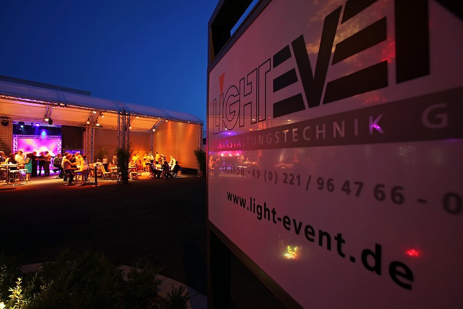 light-event Eine große Abendveranstaltung im Freien zeigt Gruppen von Menschen, die an Tischen sitzen, umgeben von bunter Bühnenbeleuchtung, sowie eine Nahaufnahme eines Schildes mit der Aufschrift „light-event.de“ im Vordergrund.