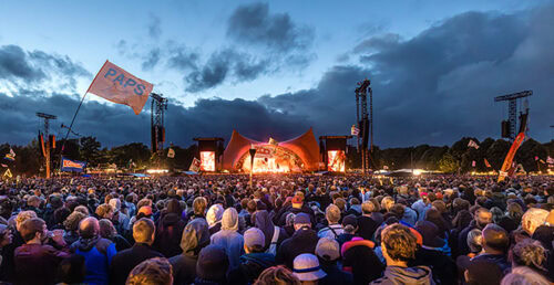 Eine große Menschenmenge besucht bei Einbruch der Dämmerung ein Open-Air-Konzert. Die Bühne ist hell beleuchtet; man sieht Zelte, Fahnen und Lautsprecher. Der Himmel ist bewölkt, und das Publikum blickt zur Bühne.