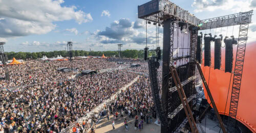 Bei einem Musikfestival versammelt sich unter teilweise bewölktem Himmel eine große Menschenmenge im Freien vor einer Bühne mit orangefarbenen Konstruktionen, Lichtern und Lautsprechern.